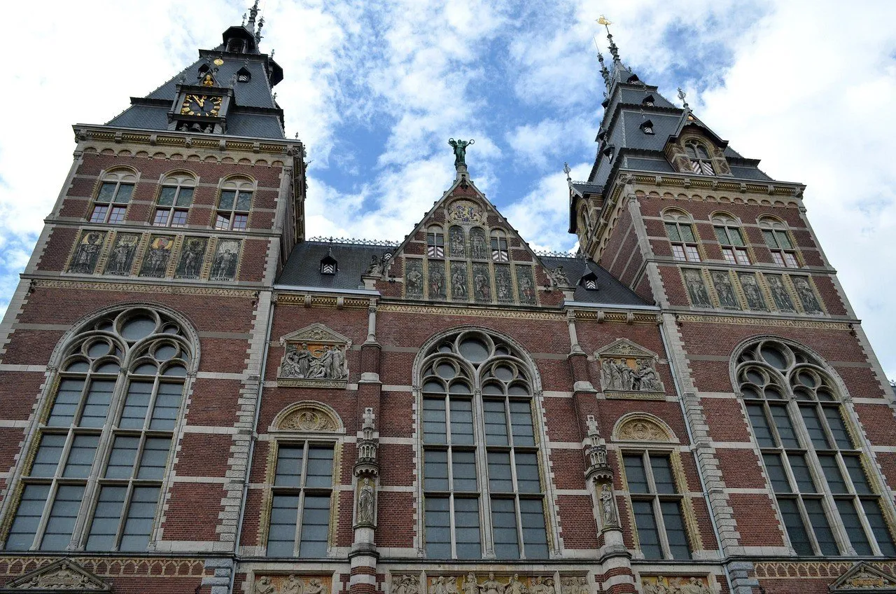 Torres, janelas e relevos da fachada de tijolos do Rijksmuseum sob céu com nuvens.