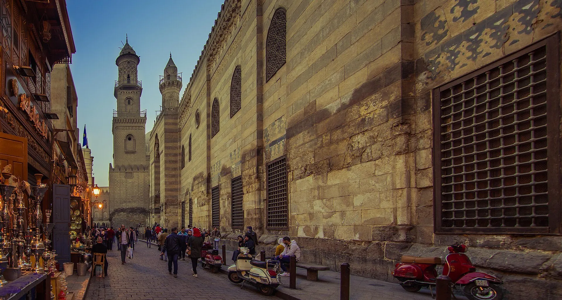 Rua Al-Muizz no Cairo, com lojas à esquerda, fachada de pedra e minaretes à direita, pedestres e scooters.