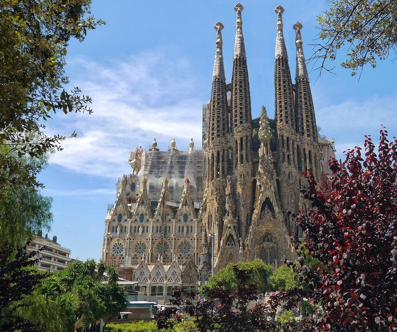 Exterior da Sagrada Família com torres da fachada da Natividade, jardins e andaimes na lateral.
