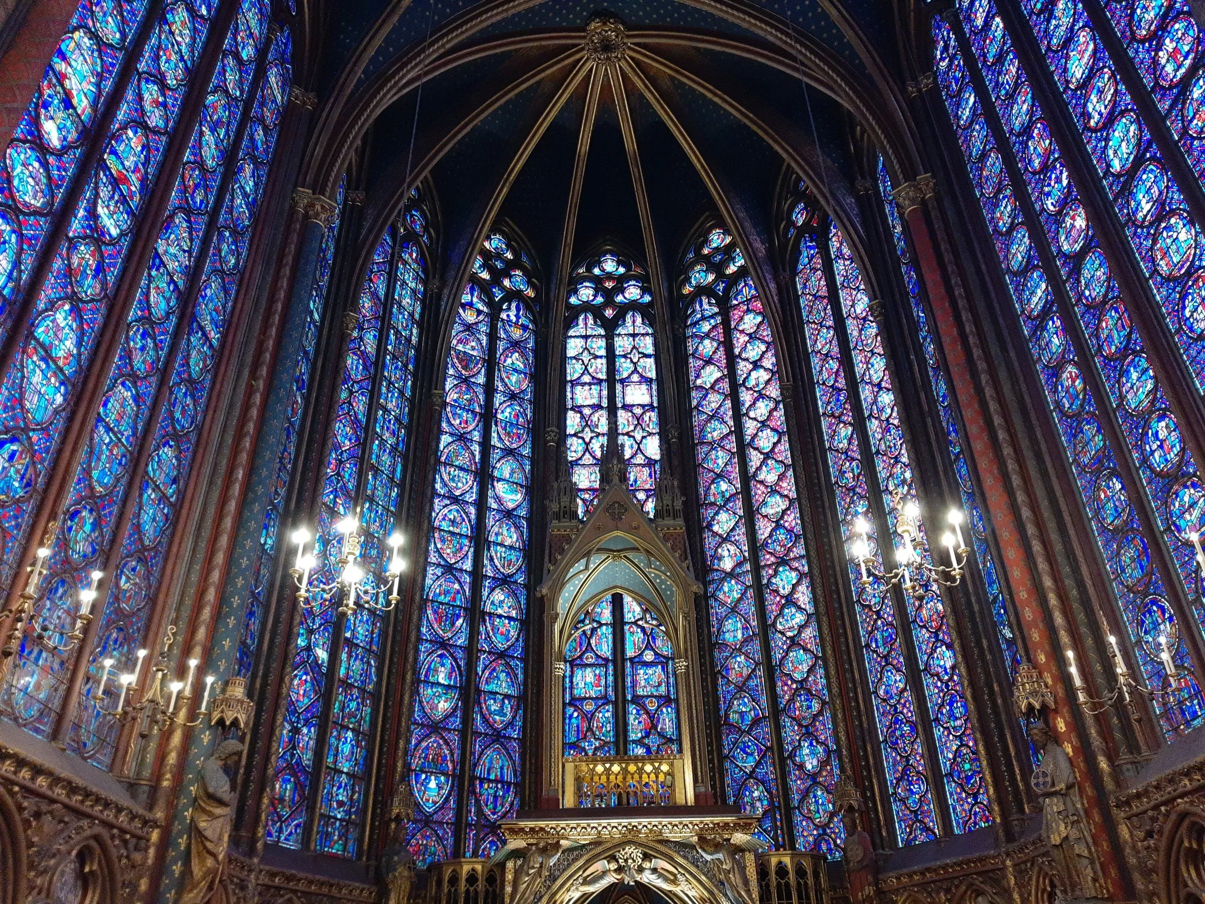 Interior da capela alta da Sainte-Chapelle, com vitrais azuis e vermelhos até a abóbada e lustres acesos ao redor do altar.