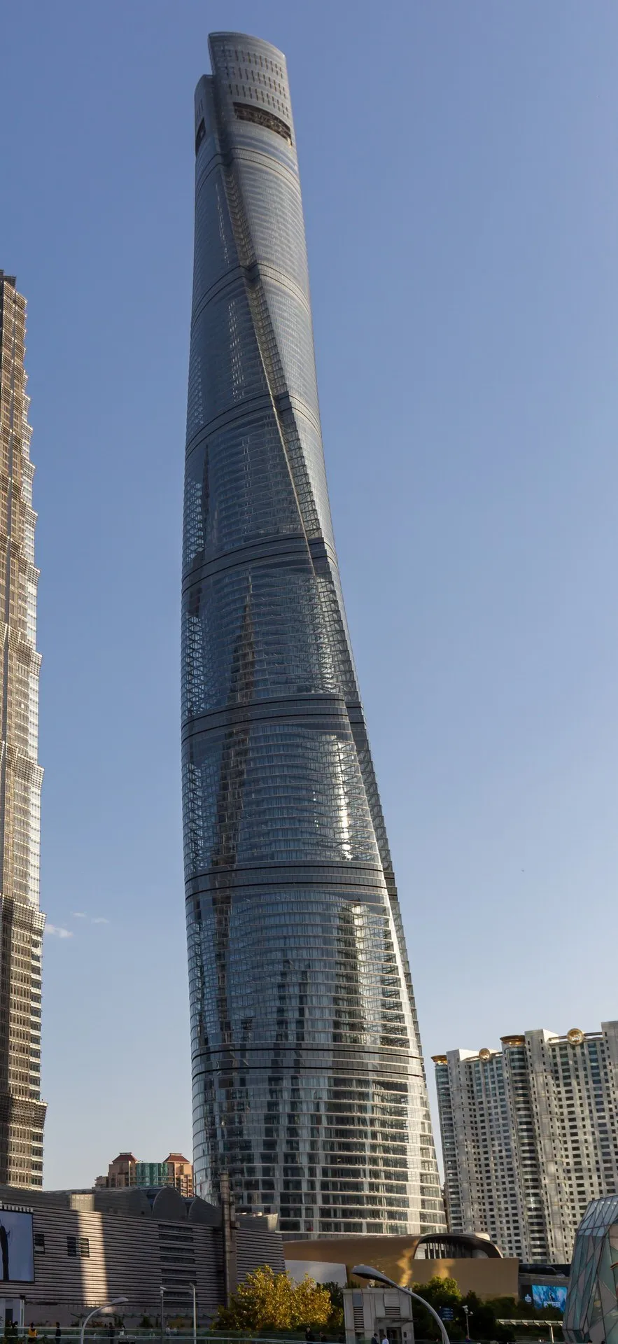 Shanghai Tower em enquadramento vertical, com fachada de vidro torcida, céu azul e prédios junto à base.