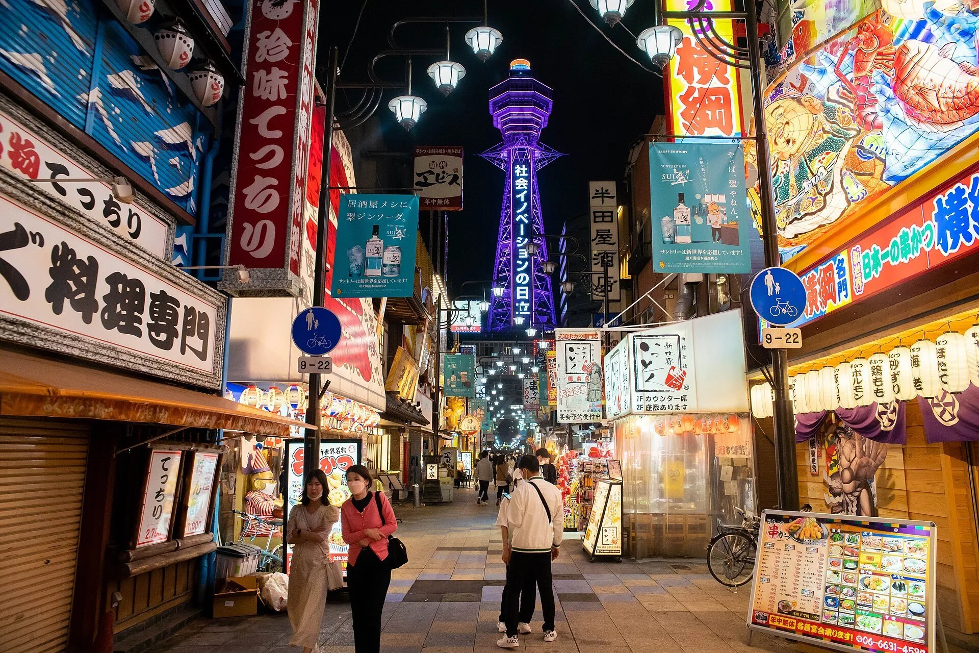 Rua de Shinsekai à noite, com restaurantes iluminados, pedestres e torre Tsutenkaku roxa ao fundo.