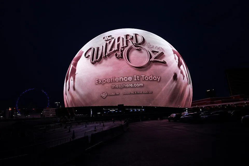 Sphere iluminada com anúncio de The Wizard of Oz na fachada, sob céu noturno, com área de acesso à frente.