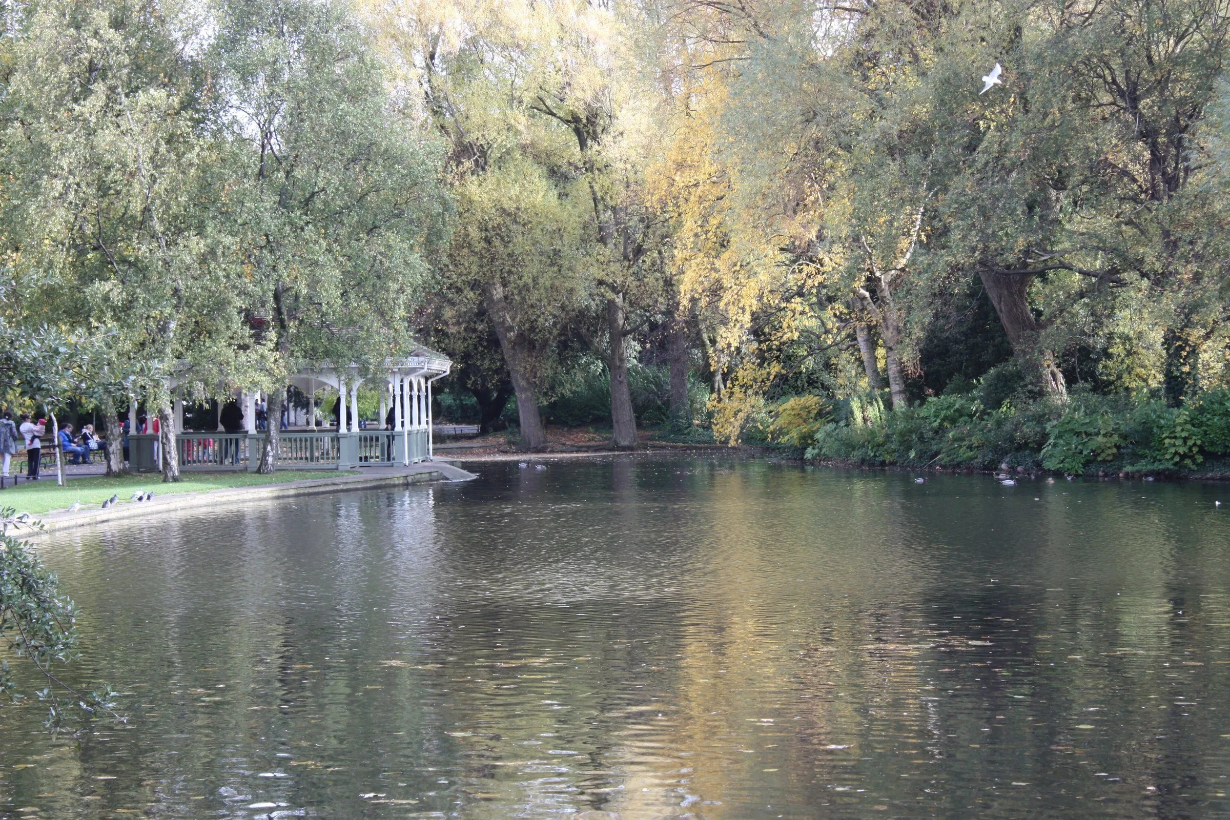 Lago de St Stephen’s Green com árvores de outono, aves na água e pavilhão branco à esquerda.