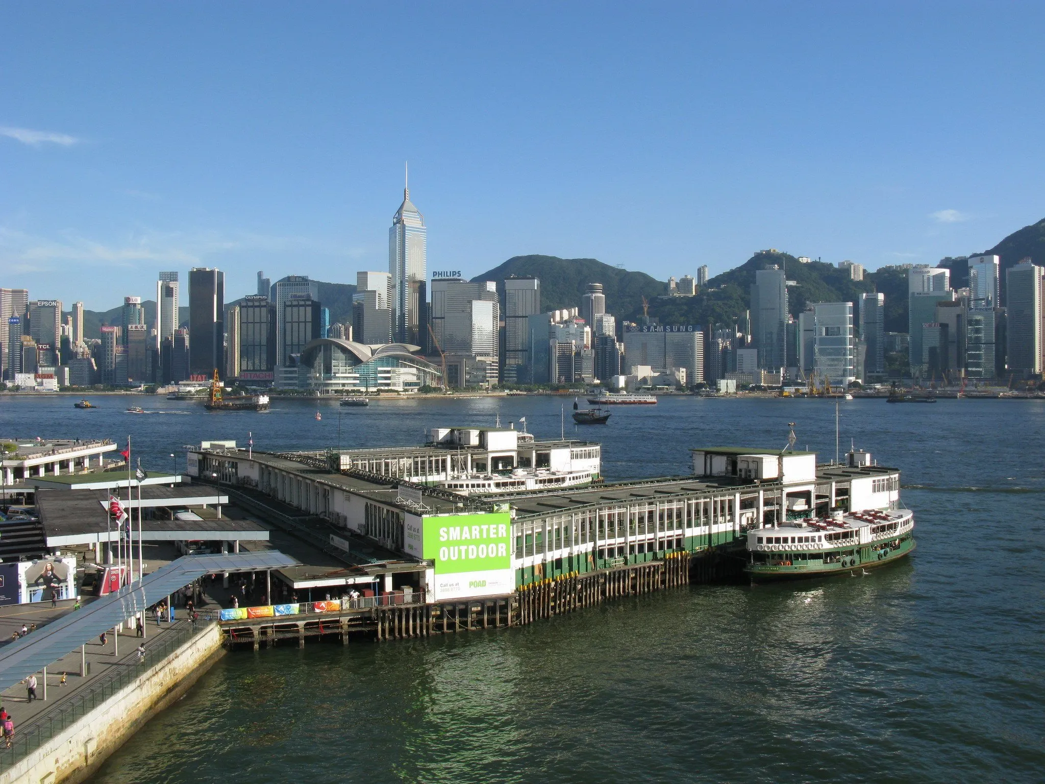 Píer do Star Ferry em Tsim Sha Tsui com uma balsa verde e branca atracada e o skyline da ilha de Hong Kong ao fundo.