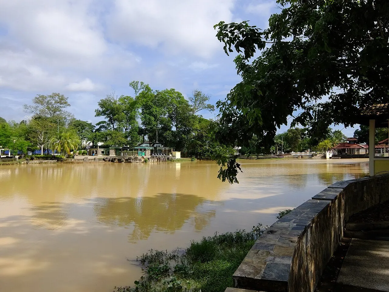 Lago de Taman Merdeka em Johor Bahru, com árvores, reflexos e caminho junto à margem.