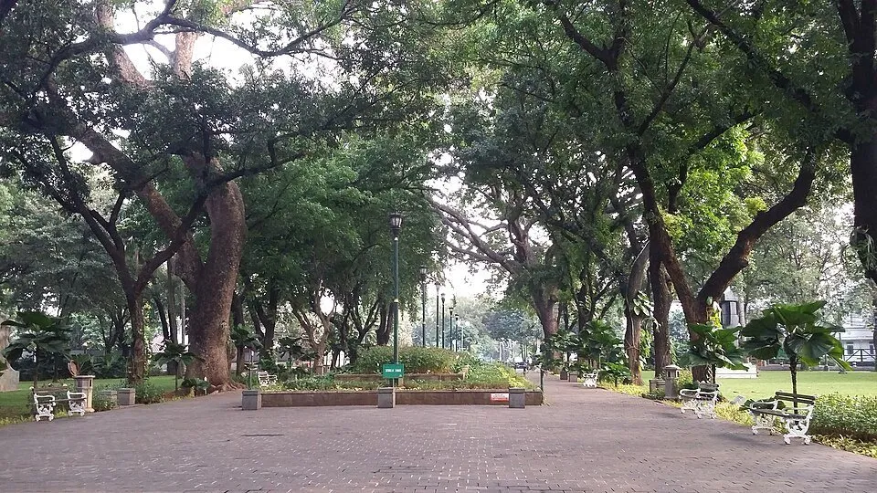 Alameda pavimentada do Taman Suropati entre árvores altas, canteiros e bancos brancos.