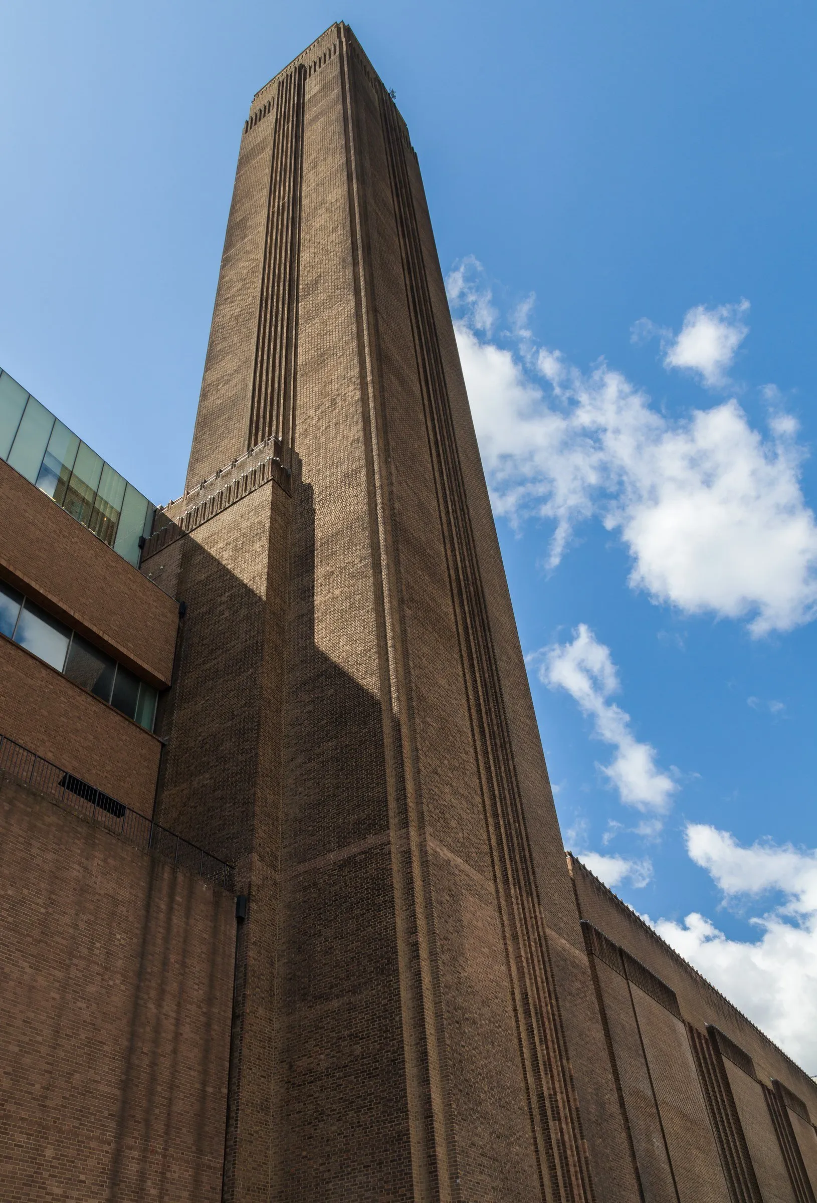 Chaminé de tijolos da Tate Modern, vista de baixo contra o céu azul com nuvens, ao lado do prédio de tijolos.