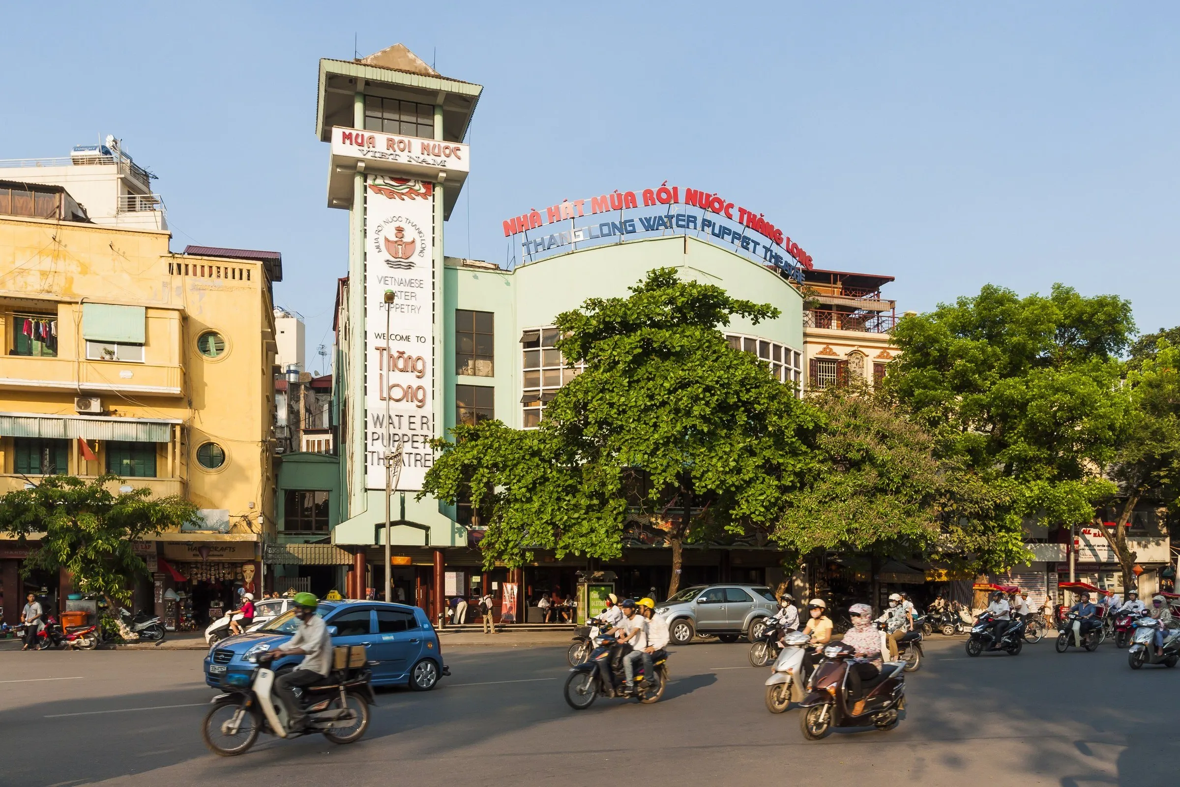 Fachada do Teatro Thang Long com letreiro de marionetes aquáticas, árvores e motos na rua.