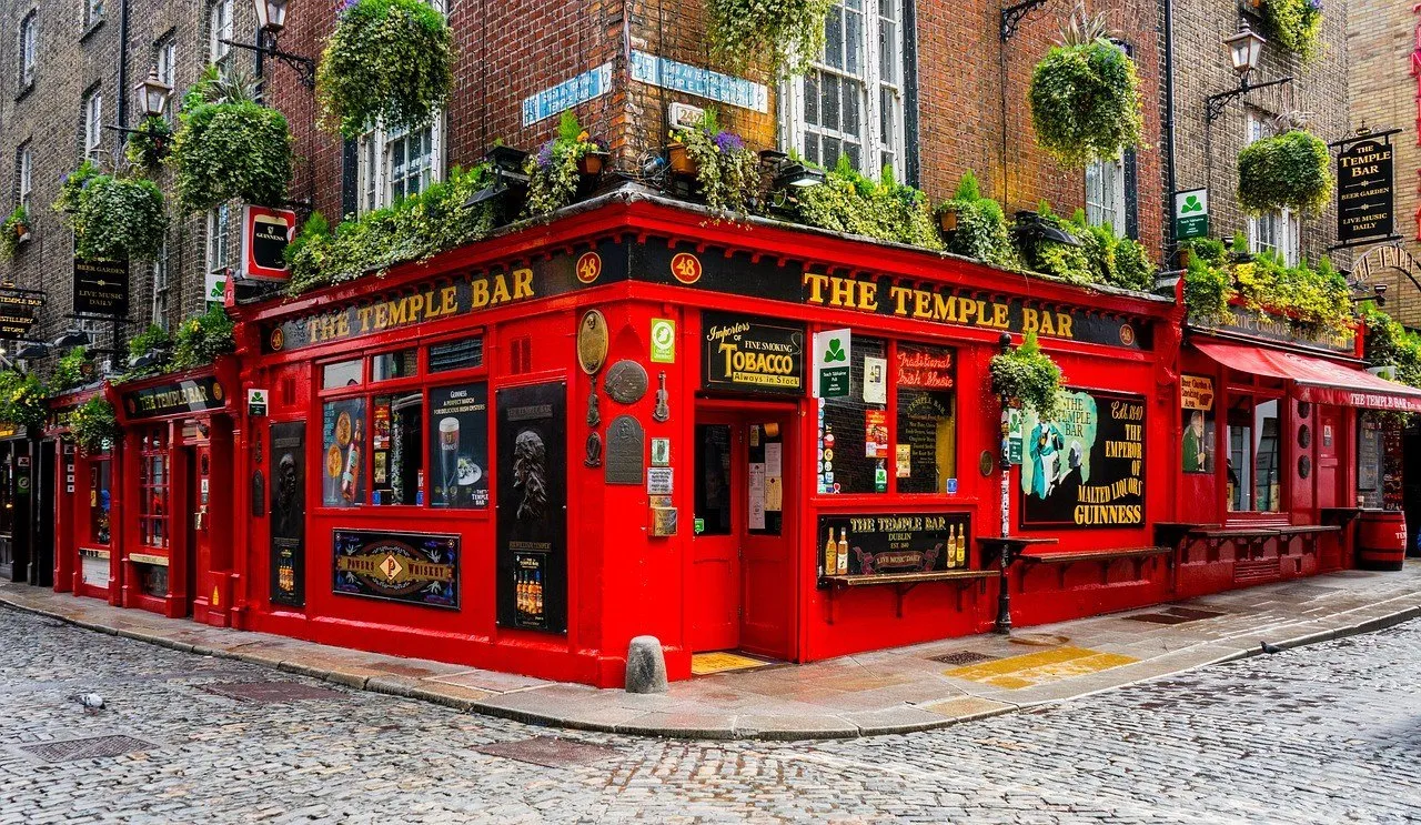 Esquina do pub The Temple Bar, com fachada vermelha, vasos de plantas e rua de pedra quase vazia.