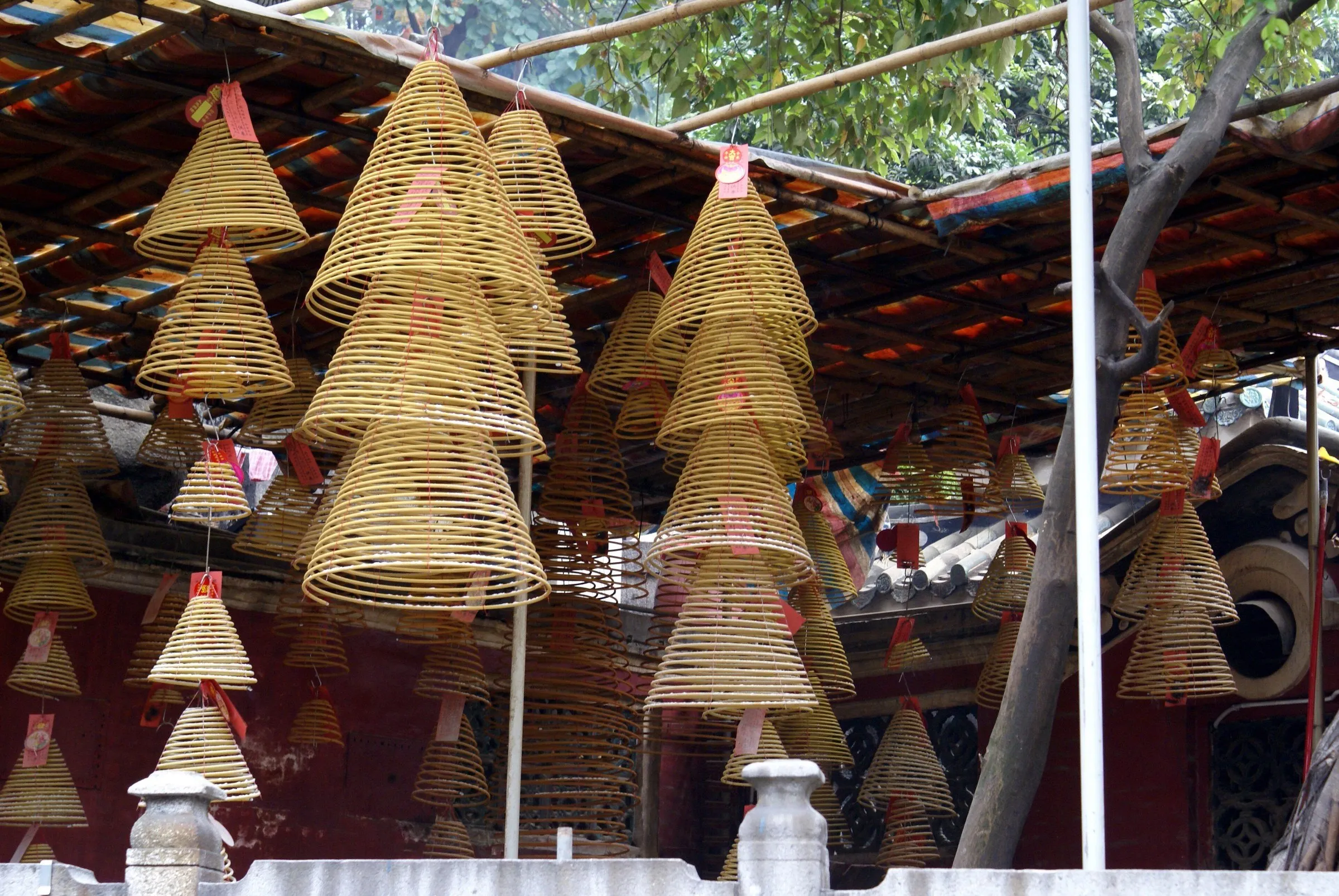 Dezenas de espirais de incenso em forma de cone penduradas sob um telhado, com muros vermelhos do templo ao fundo.