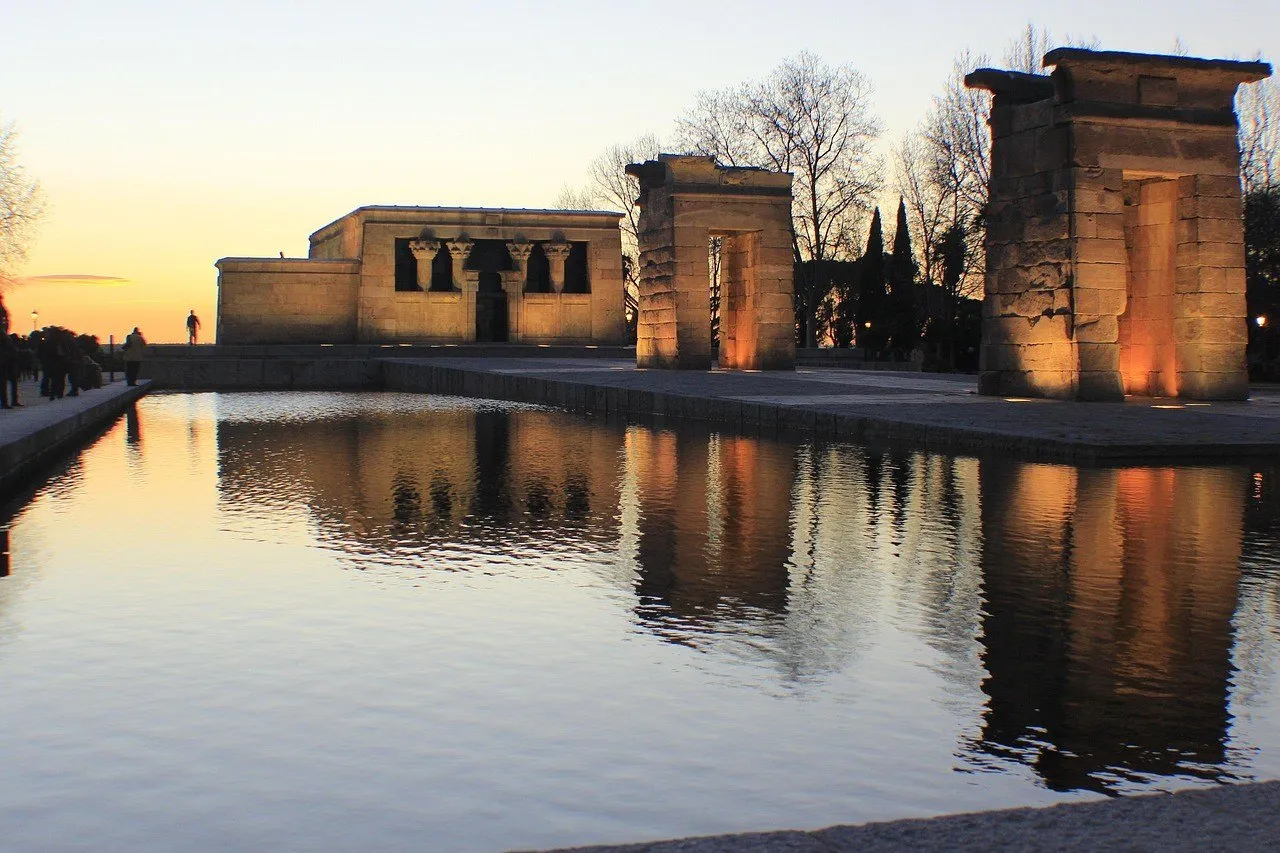 Templo de Debod e dois portais de pedra iluminados ao entardecer, refletidos na água.