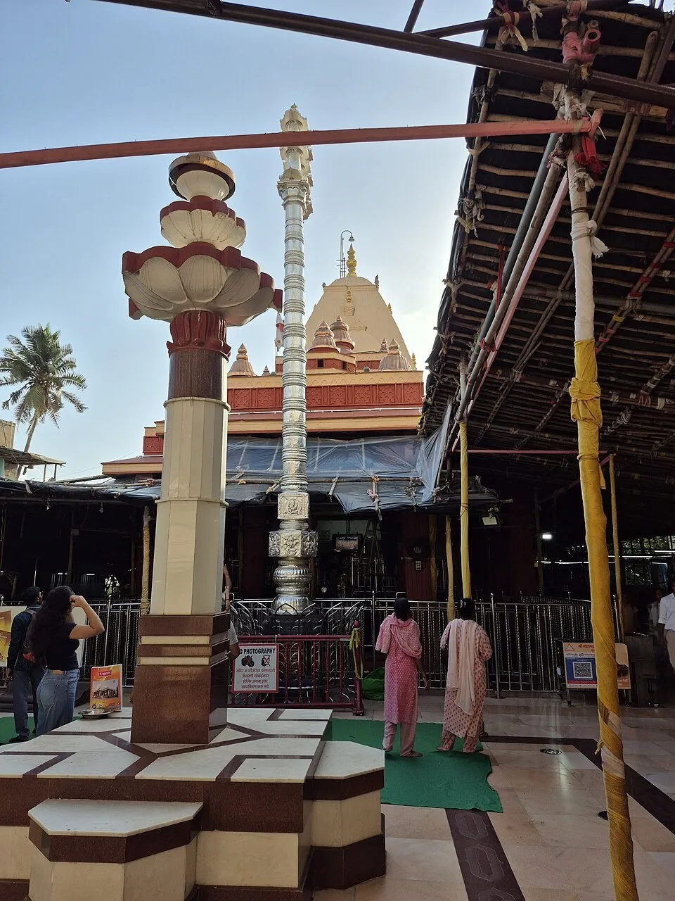Pátio do Templo de Mahalaxmi, em Mumbai, com torres amarelas e vermelhas, coluna metálica e visitantes.