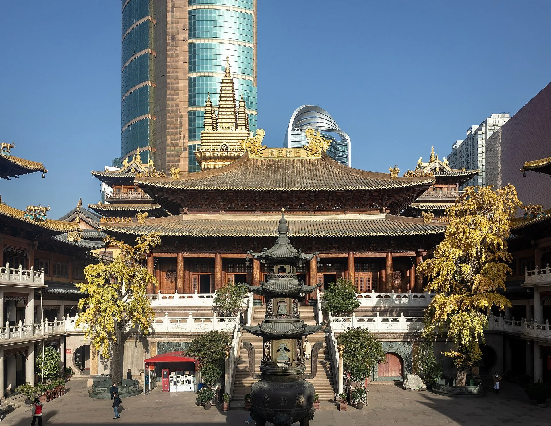 Pátio do Templo Jing’an com incensário, árvores de folhas amarelas e telhados dourados diante de prédios altos.