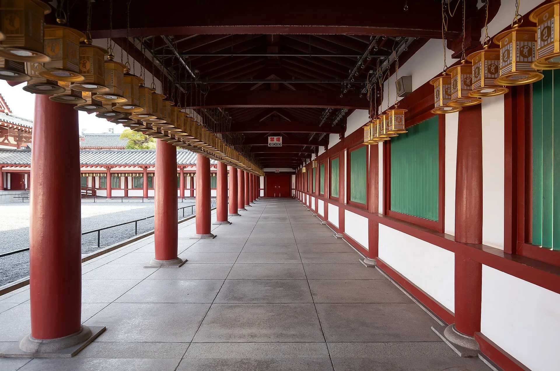 Corredor de Shitennoji com colunas vermelhas, janelas verdes e fileiras de lanternas douradas.