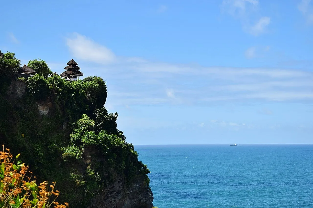 Templo de Uluwatu no alto de uma falésia coberta de vegetação, diante do oceano azul e de céu claro.