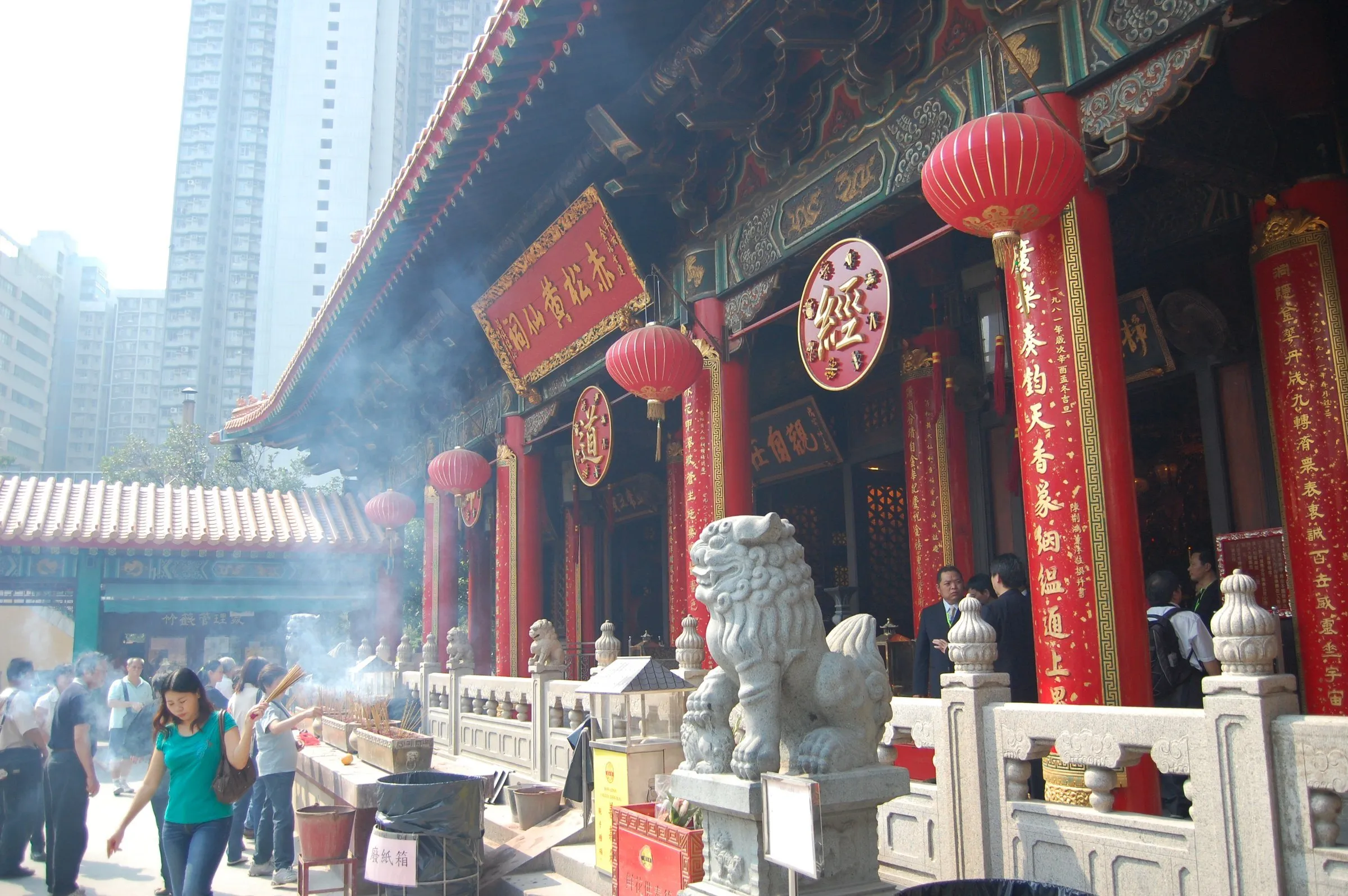 Salão principal do Templo Wong Tai Sin, com colunas vermelhas, leão de pedra, fumaça de incenso e prédios altos ao fundo.