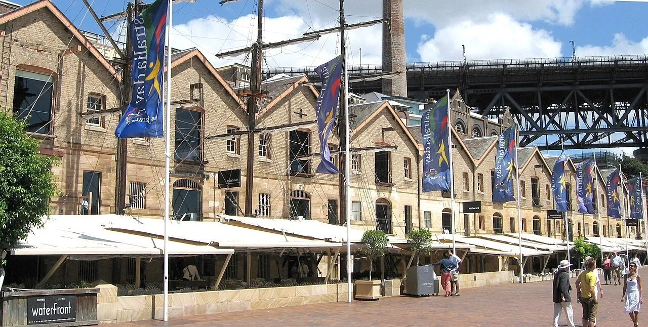 Fachadas de pedra em The Rocks, com mastros, bandeiras, mesas sob toldos e Harbour Bridge ao fundo.