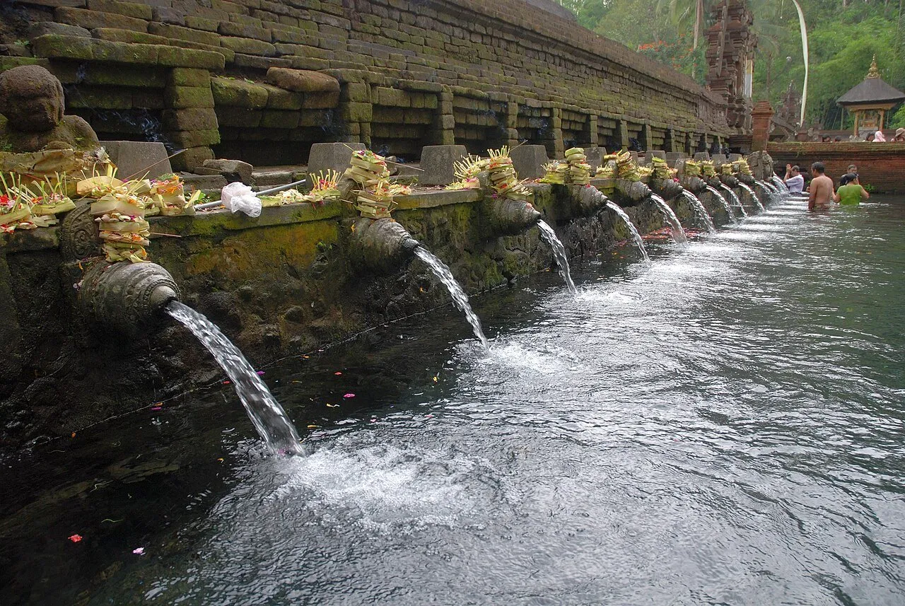 Sequência de bicas de pedra despejando água em uma piscina de Tirta Empul, com oferendas e fiéis ao fundo.