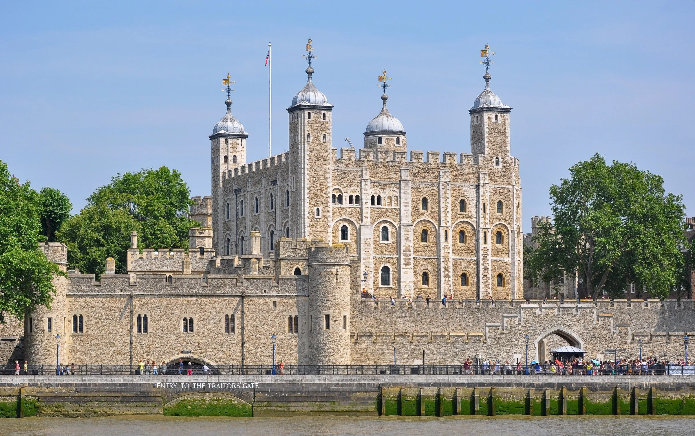 Torre Branca e muralhas de pedra da Torre de Londres, vistas da margem do Tâmisa sob céu azul.