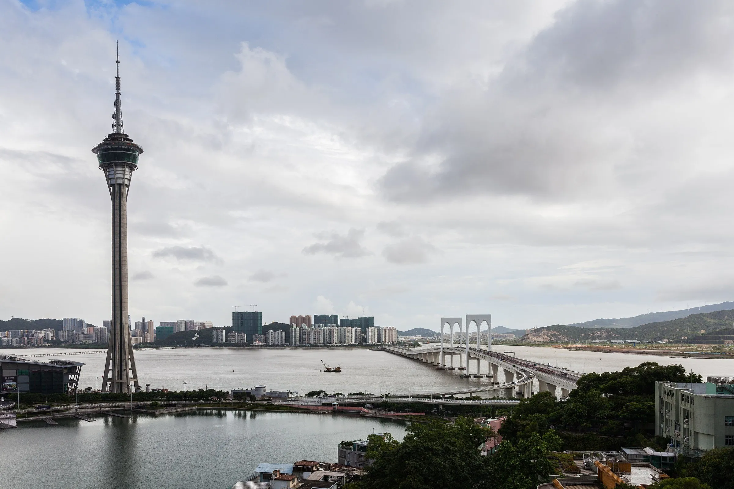 Torre de Macau vista do outro lado da água, com a baía, uma ponte em arco e a cidade ao fundo sob céu nublado.