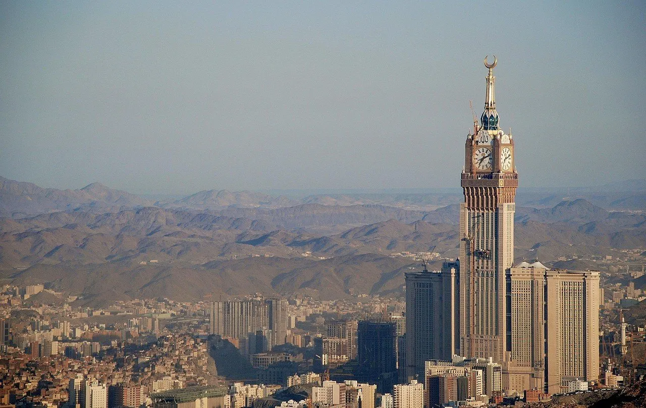 Torre do Relógio de Meca vista à distância, com edifícios abaixo e montanhas ao fundo.