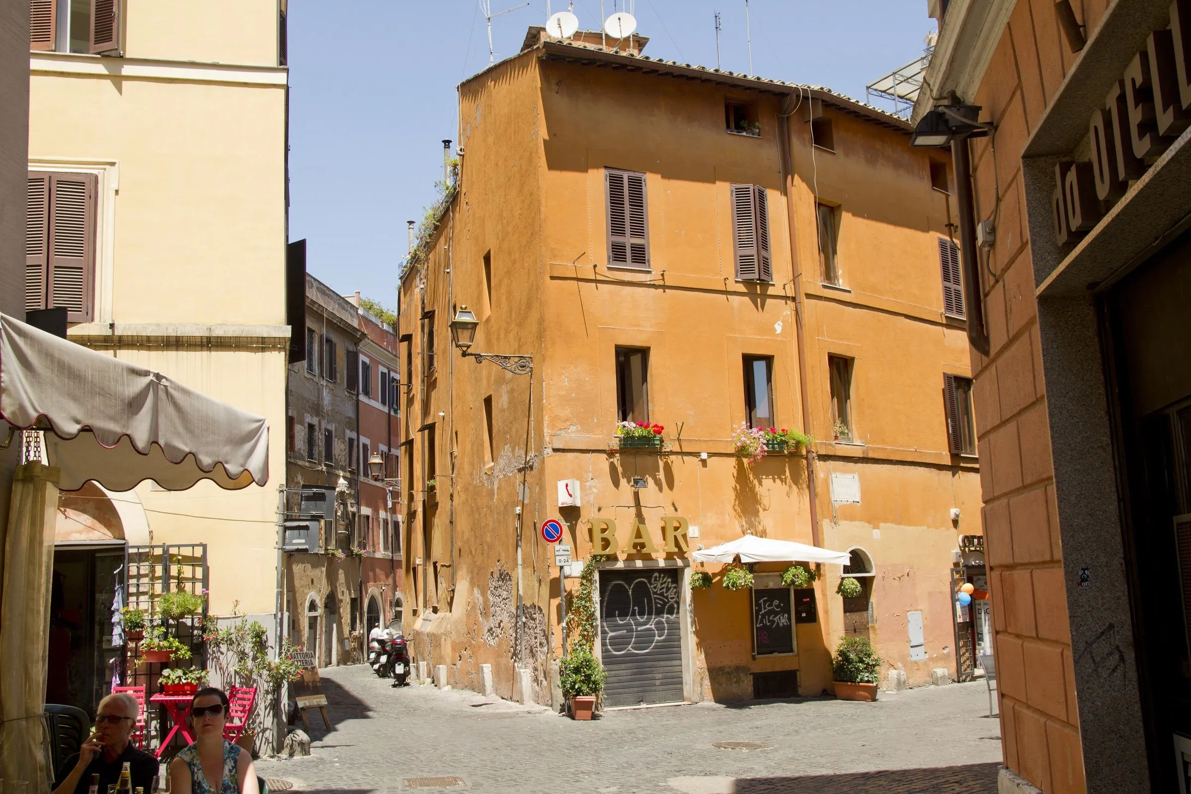 Esquina de Trastevere com casarão ocre, janelas com venezianas, mesas de um bar na calçada e rua de pedra estreita.