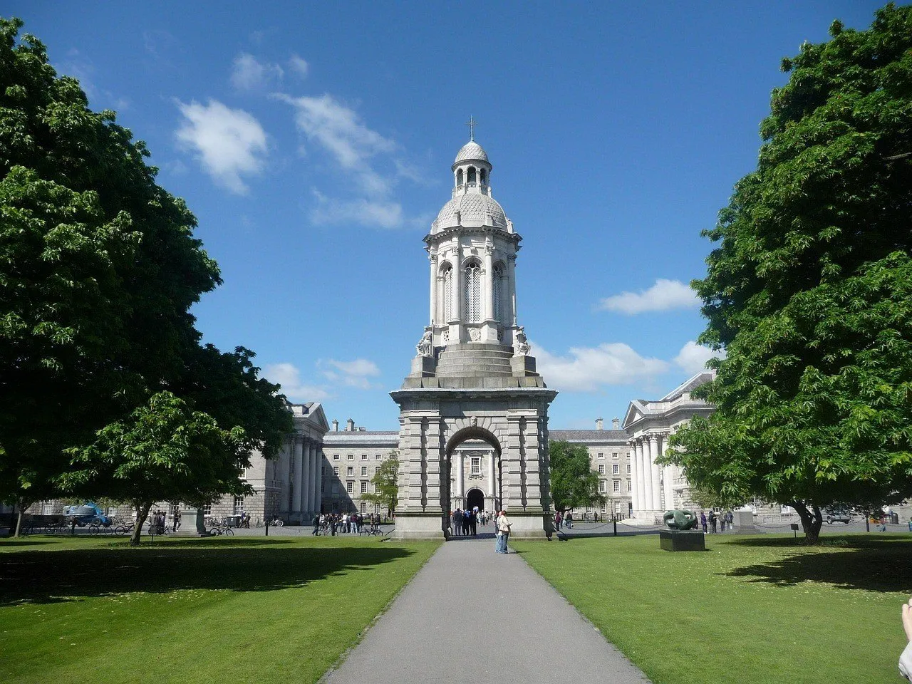 Campanário de pedra do Trinity College entre árvores, gramados e edifícios do campus, sob céu azul.