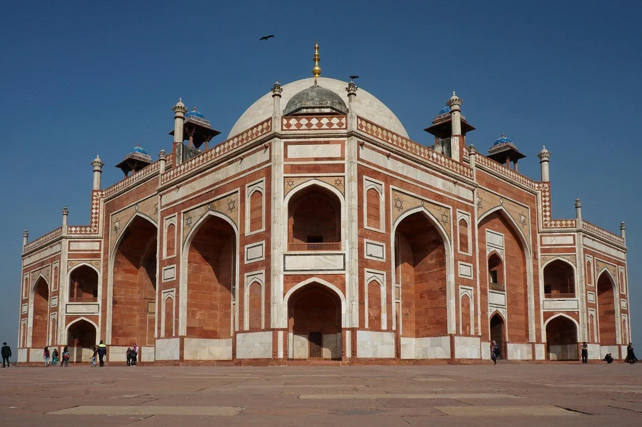 Fachada do Túmulo de Humayun, em Délhi, com arcos de arenito vermelho, frisos brancos e cúpula branca sob céu azul intenso.