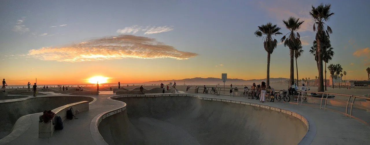 Skatepark de Venice Beach ao pôr do sol, com palmeiras, praia e visitantes ao fundo.