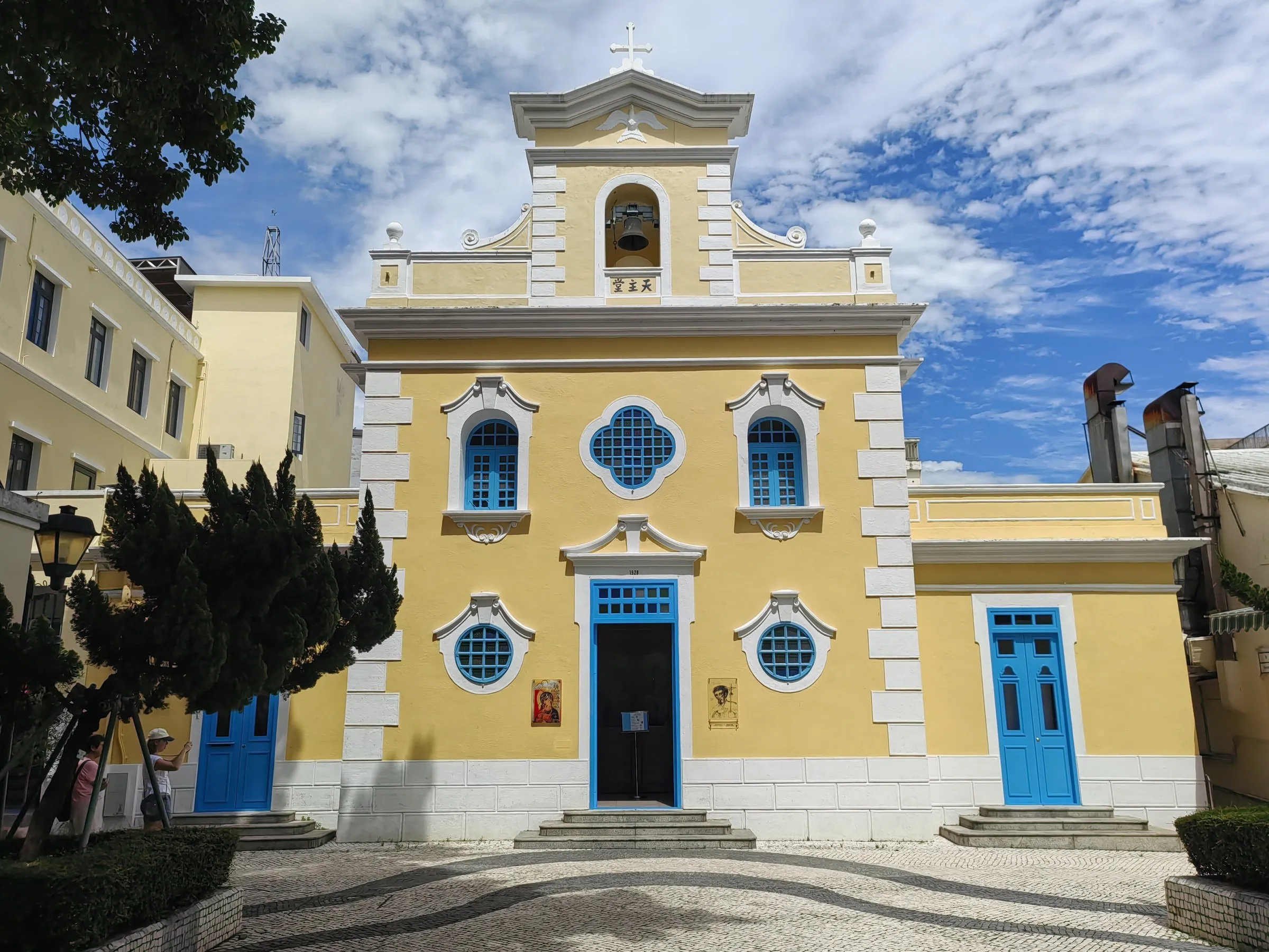 Fachada amarela e branca da Capela de São Francisco Xavier, com portas azuis, em um largo de calçada ondulada sob céu azul.