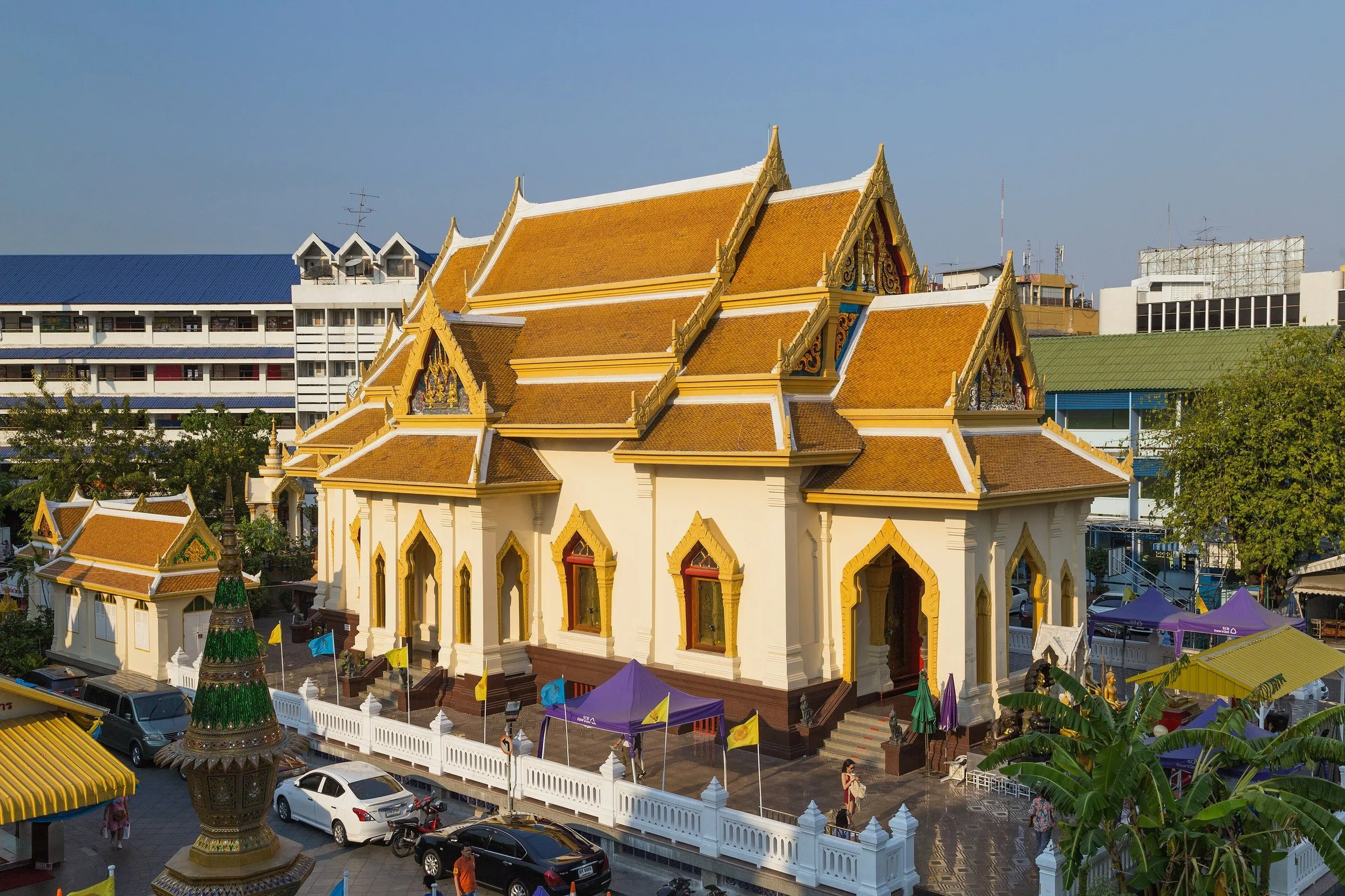 Salão de telhado dourado e paredes creme do Wat Traimit, em Bangkok, visto de cima, com carros e barracas ao redor.