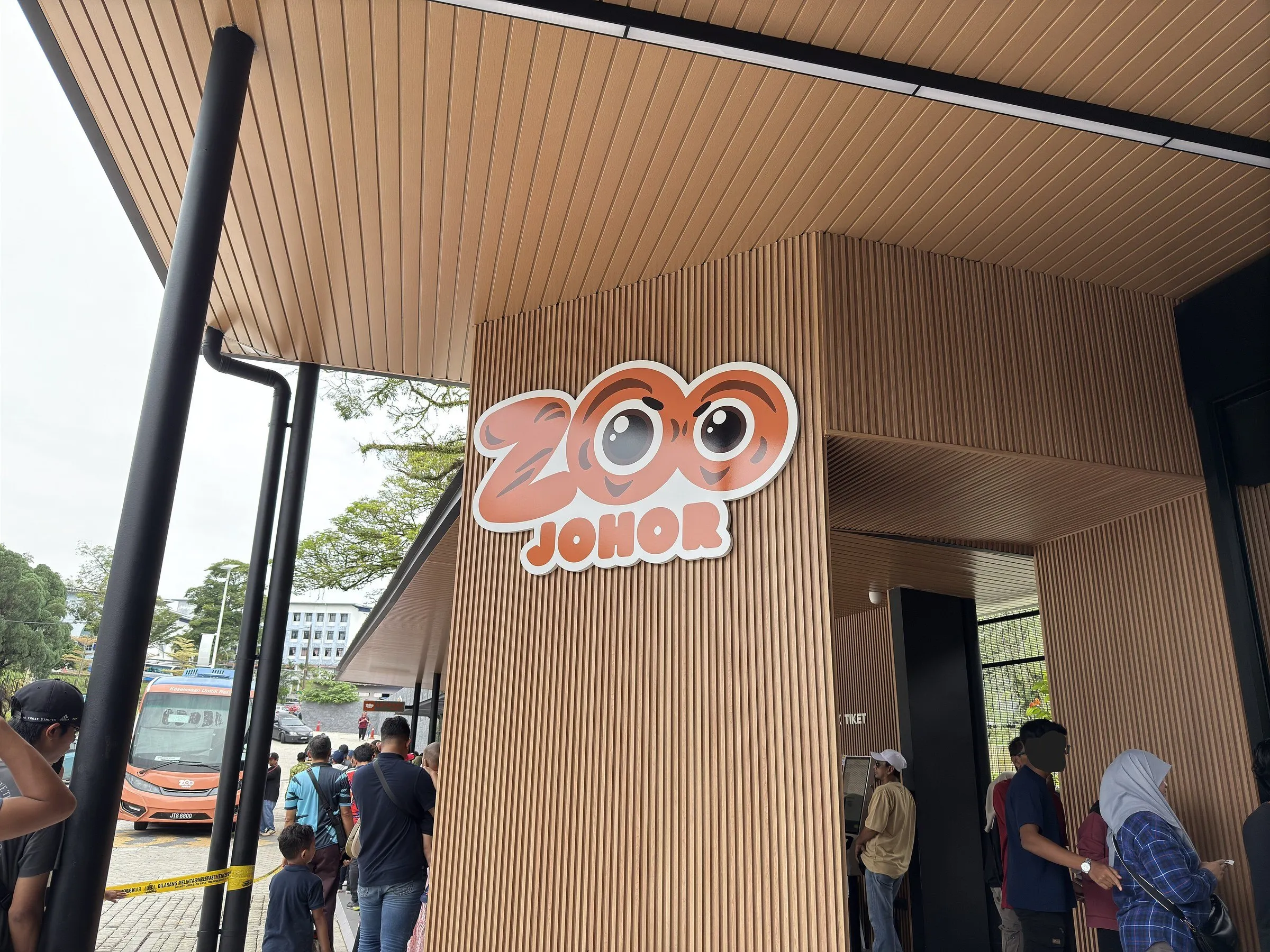 Entrada do Zoo Johor com fachada de ripas, letreiro ilustrado e visitantes junto à bilheteria.